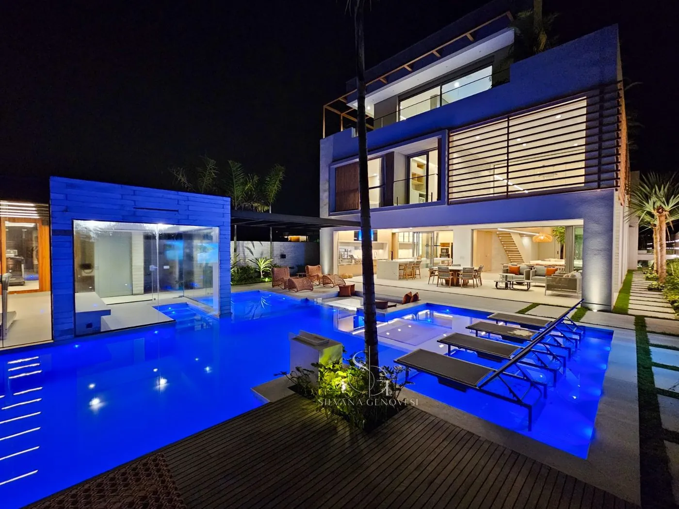 Fachada e piscina iluminadas à noite, com espreguiçadeiras e living aberto