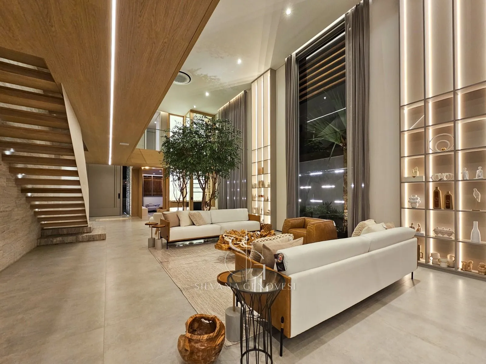 Living com árvore interna, sofás claros, poltrona de couro e estante iluminada