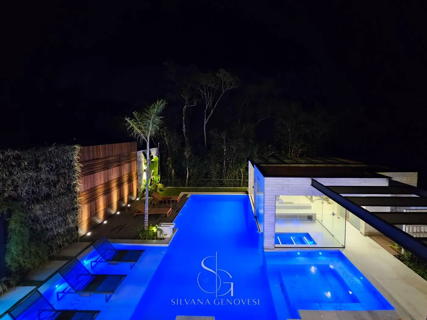 Piscina iluminada à noite vista do alto, com deck, painel de madeira e mata ao fundo